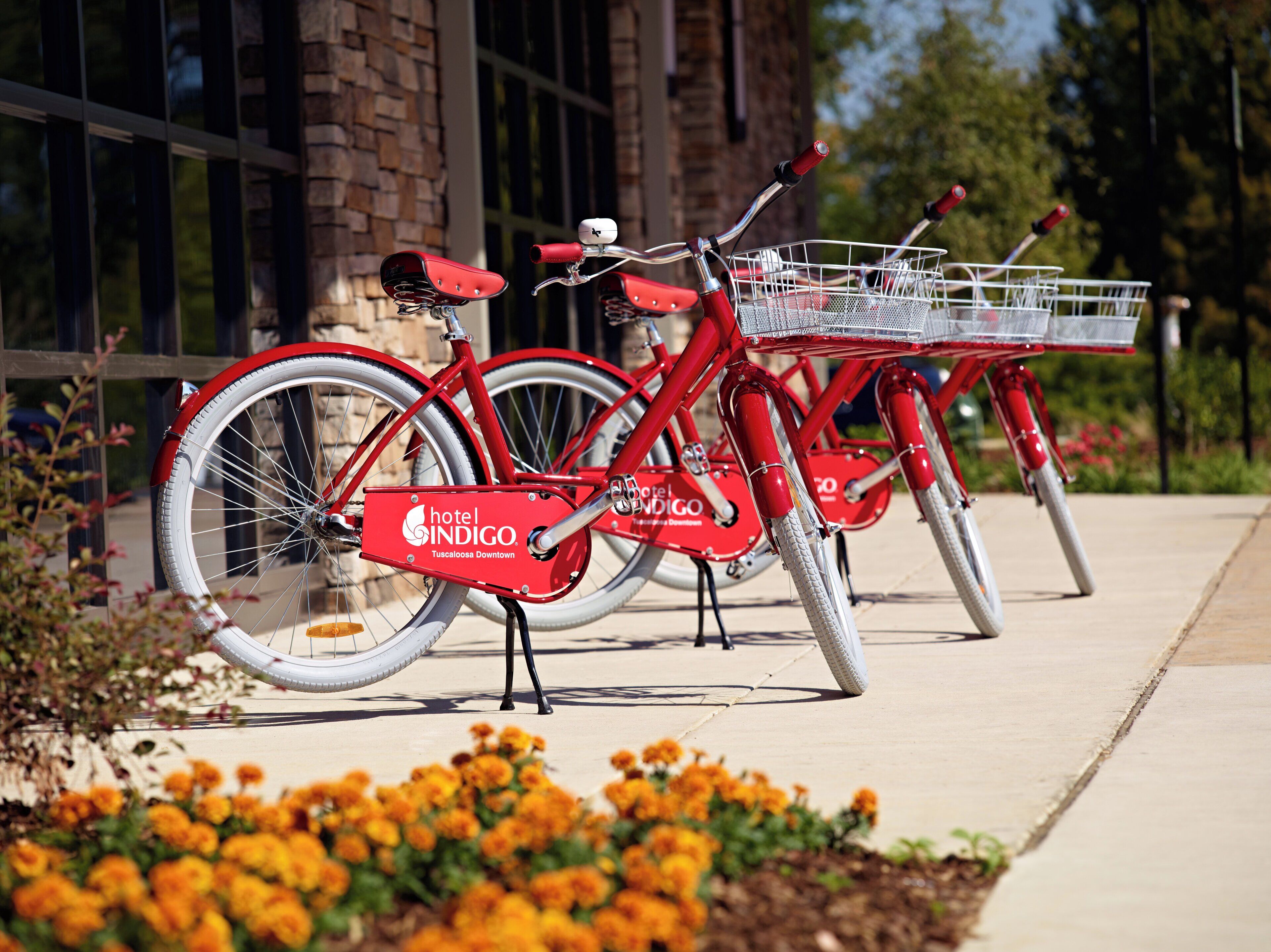 Una bicicleta roja con canasta, estacionada frente a un edificio con un letrero que dice 'hotel INDIGO Tuscaloosa Downtown'.