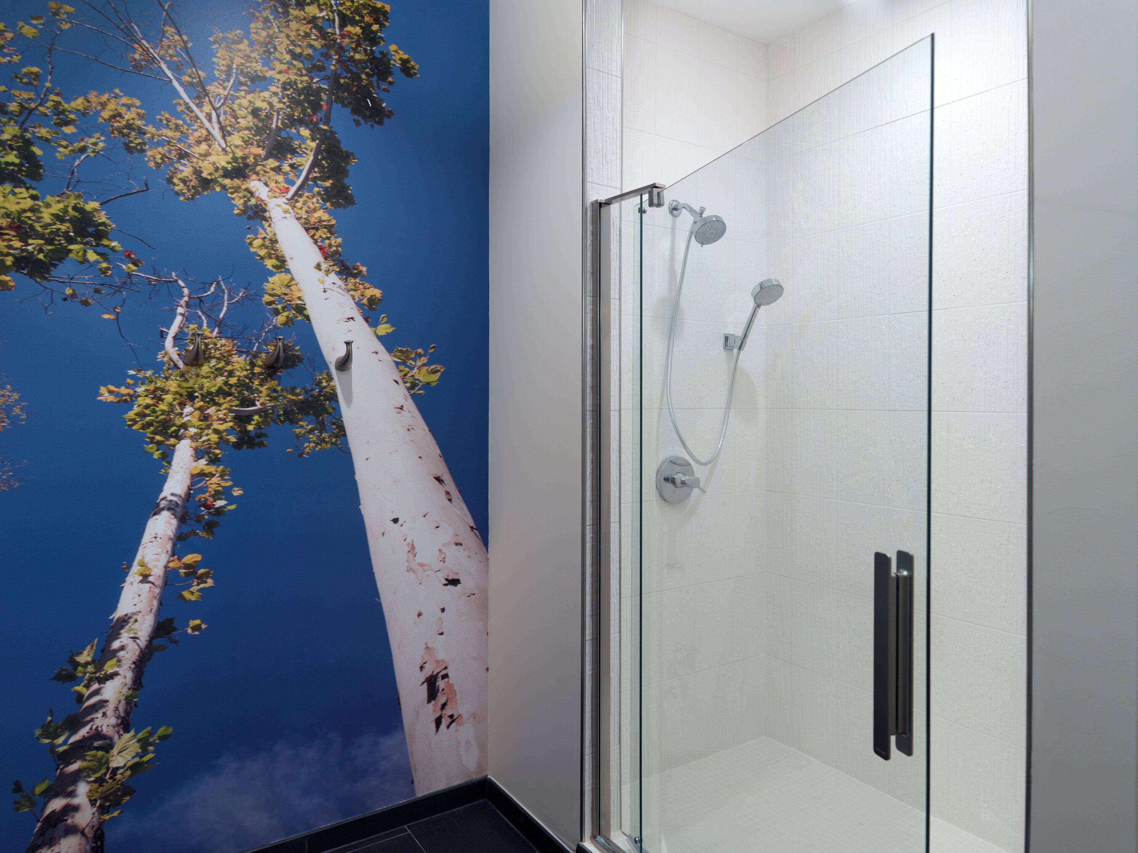 A glass-enclosed shower with a modern design, a wall with a tree mural, and a clean, white tiled floor.