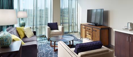 A modern hotel room with a sofa, armchair, coffee table, and a TV on a wooden cabinet.