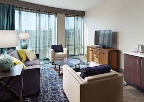 Quarto de hotel moderno com sofá, poltrona, mesa de centro e TV sobre um móvel de madeira.