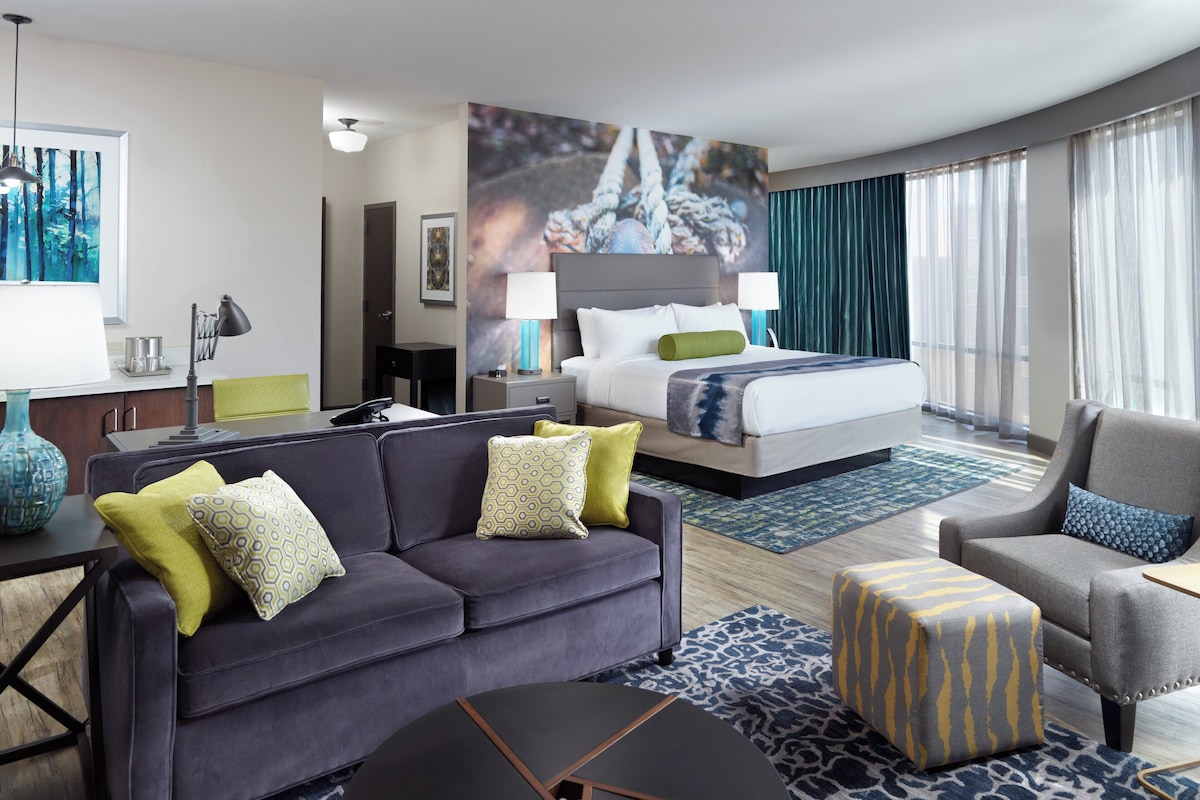 Quarto de hotel moderno com uma cama grande, um sofá, uma poltrona, uma mesa de escritório e uma mesa de centro.