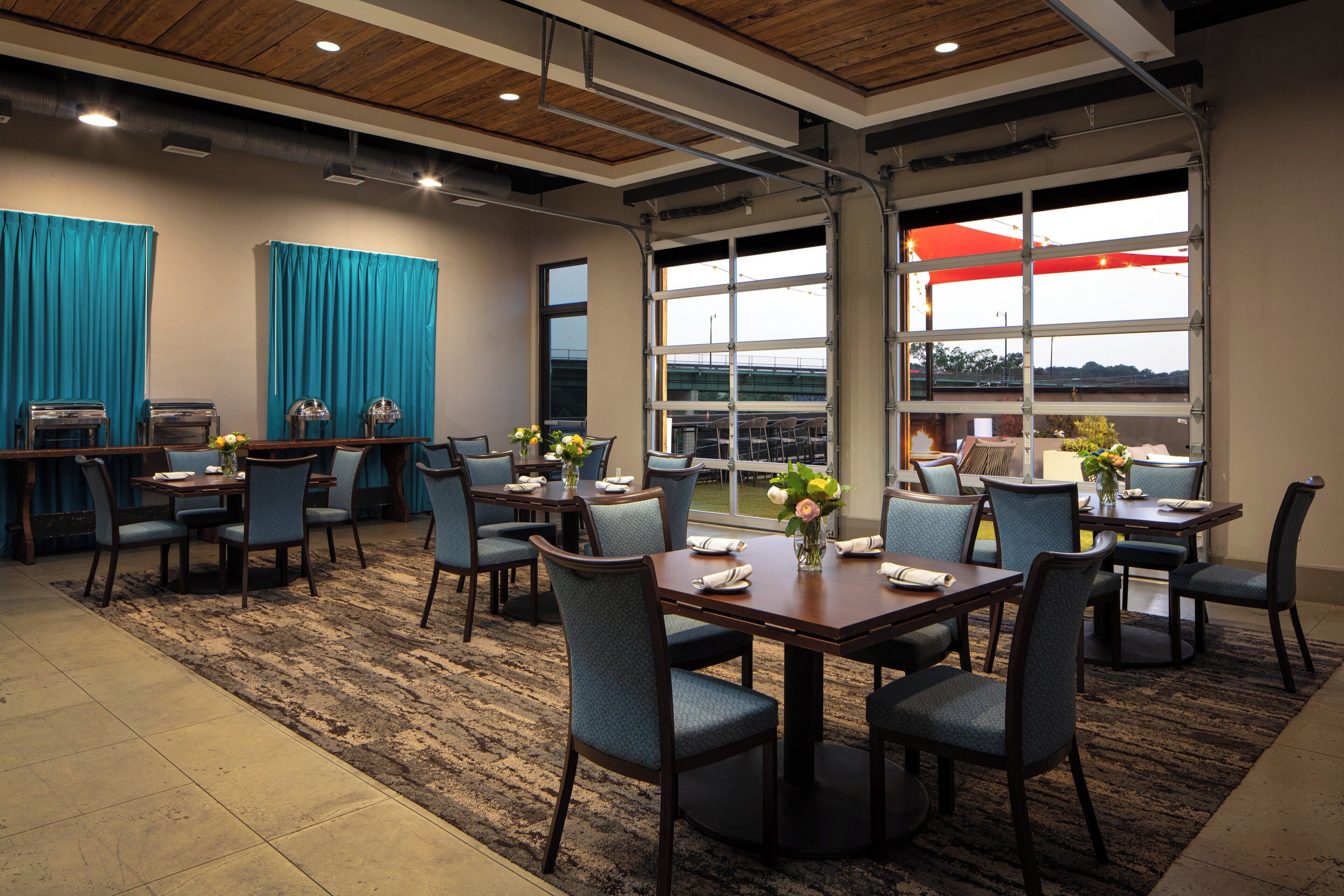A hotel dining area with multiple tables and chairs, large windows, and a view of the outdoors.
