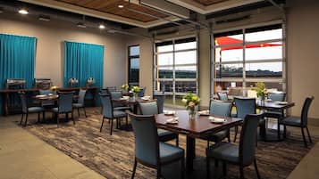 A hotel dining area with multiple tables and chairs, large windows, and a view of the outdoors.