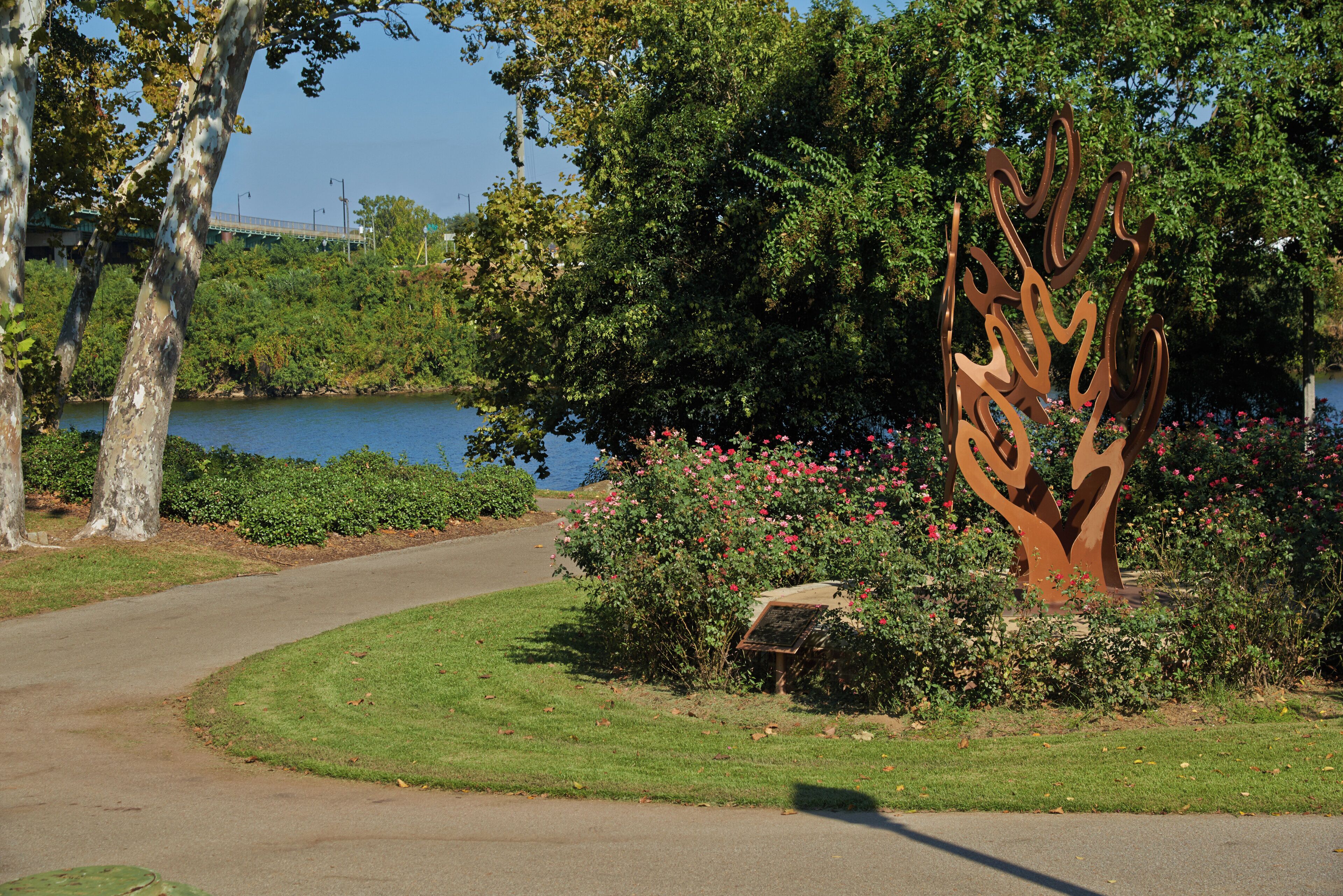 A park with a curved pathway, a metal sculpture, and a body of water in the background.