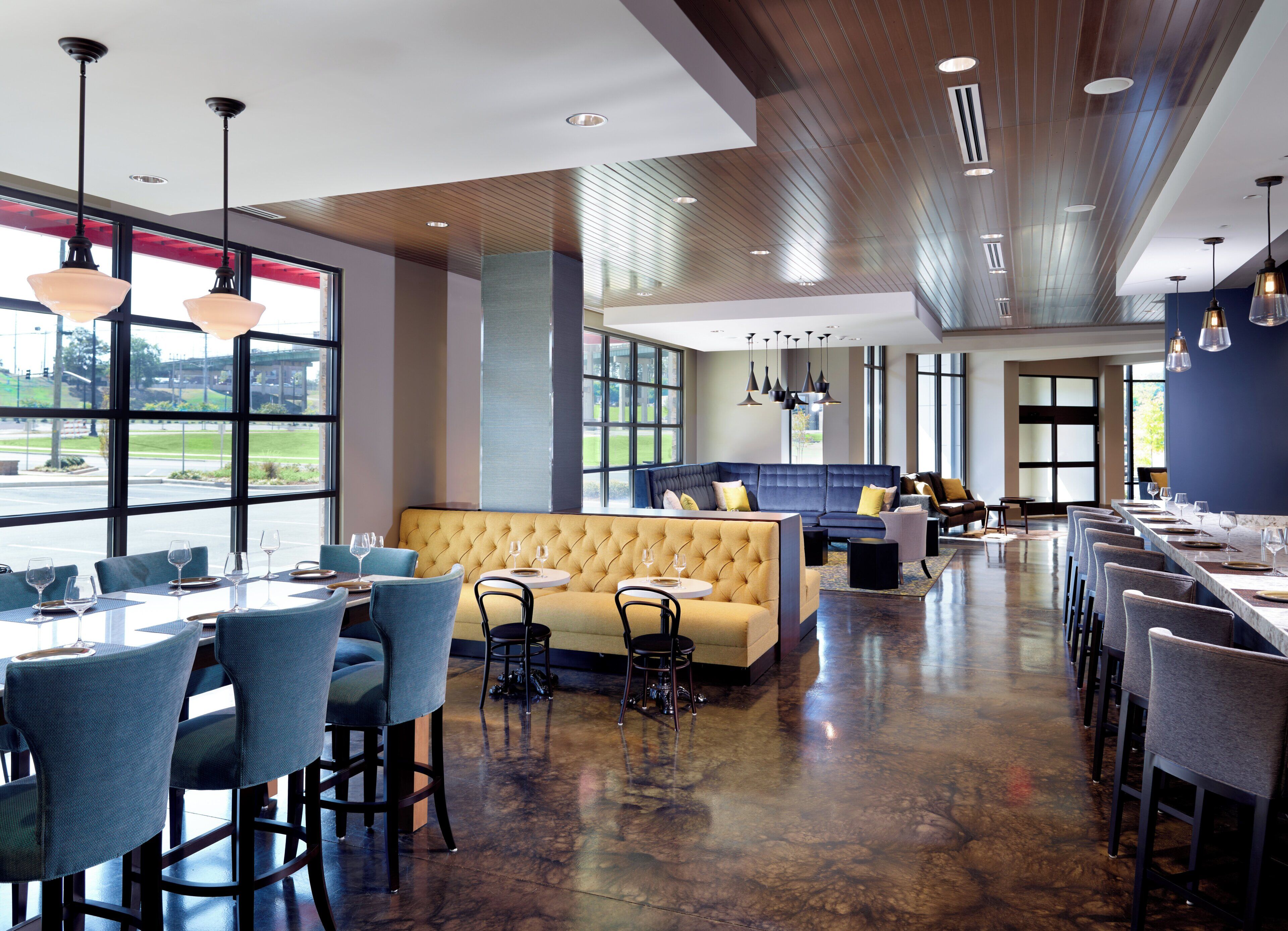 A spacious restaurant with a long counter, multiple seating areas including a sofa and chairs, and large windows offering a view of the outdoors.