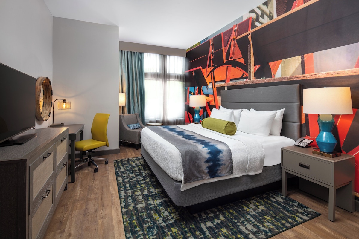 Quarto de hotel com cama, mesas de cabeceira, escrivaninha, cadeira, televisão e um grande quadro abstrato na parede.