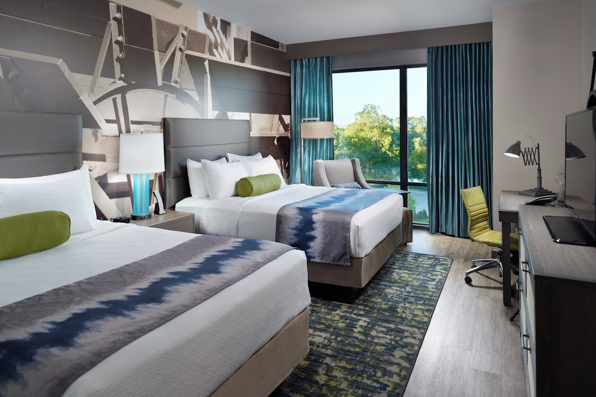 Quarto de hotel com duas camas, uma escrivaninha, uma cadeira e uma janela grande com vista para árvores.