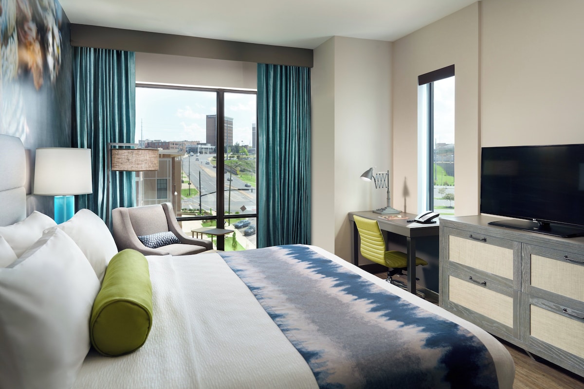 Quarto de hotel com cama, escrivaninha, cadeira, televisão e vista para a paisagem urbana.