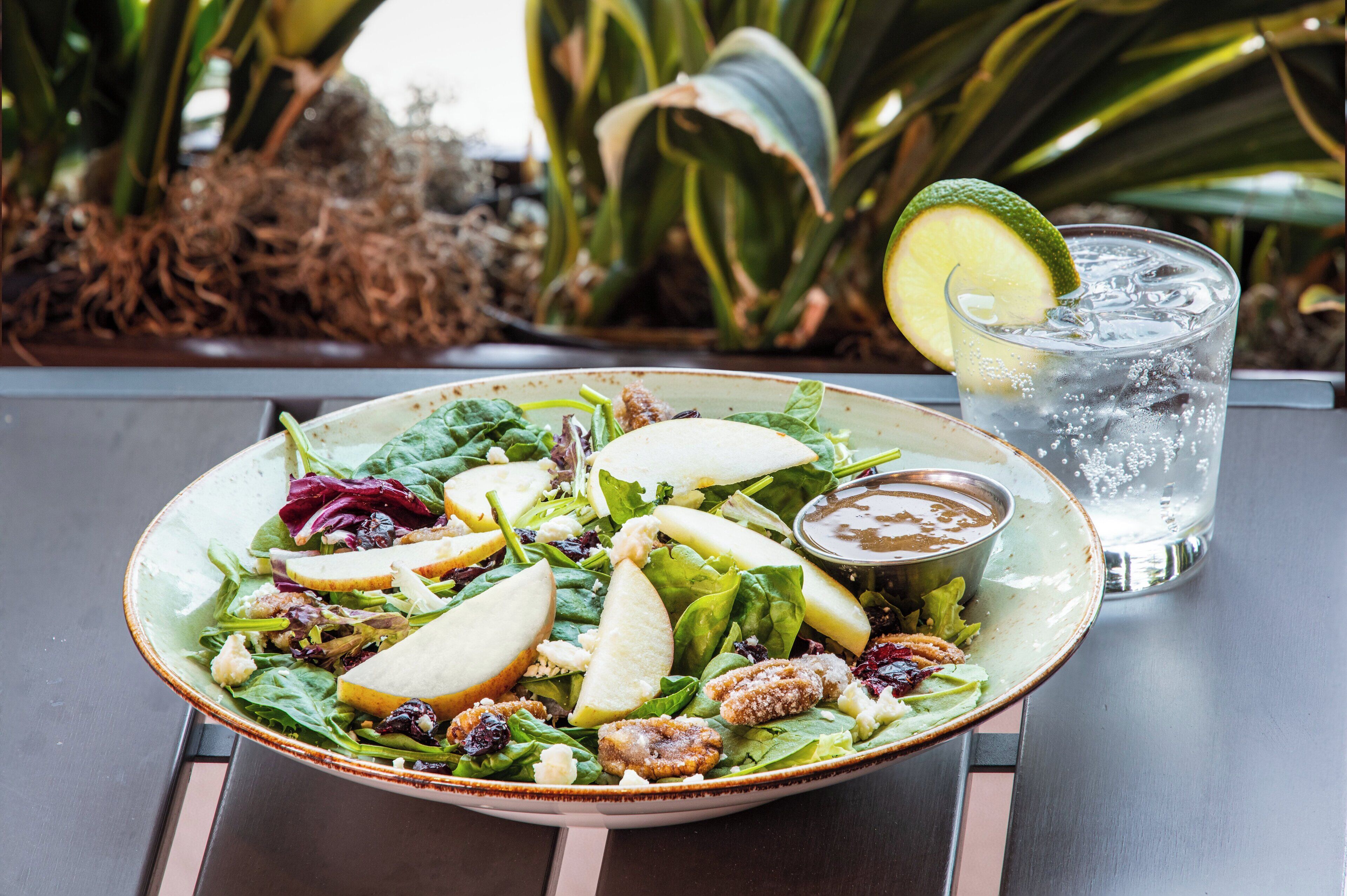 Un saladier rempli de salade, avec des pommes, des noix et un quartier de citron vert, posé sur une table à côté d’un verre d’eau.