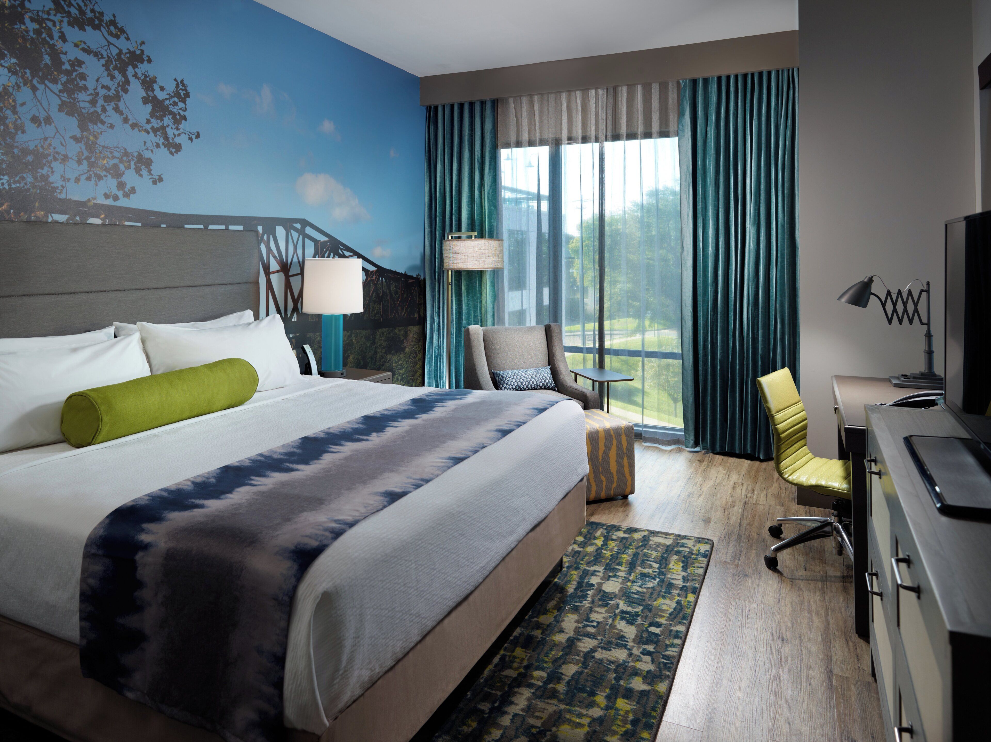 A hotel room with a bed, a desk, a chair, a TV, a lamp, and a view of greenery through the window.