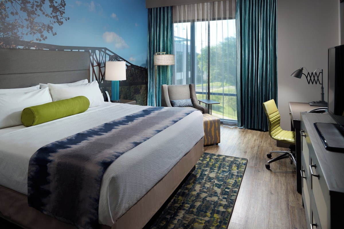 Quarto de hotel com cama, escrivaninha, cadeira, televisão, abajur e vista para o verde pela janela.