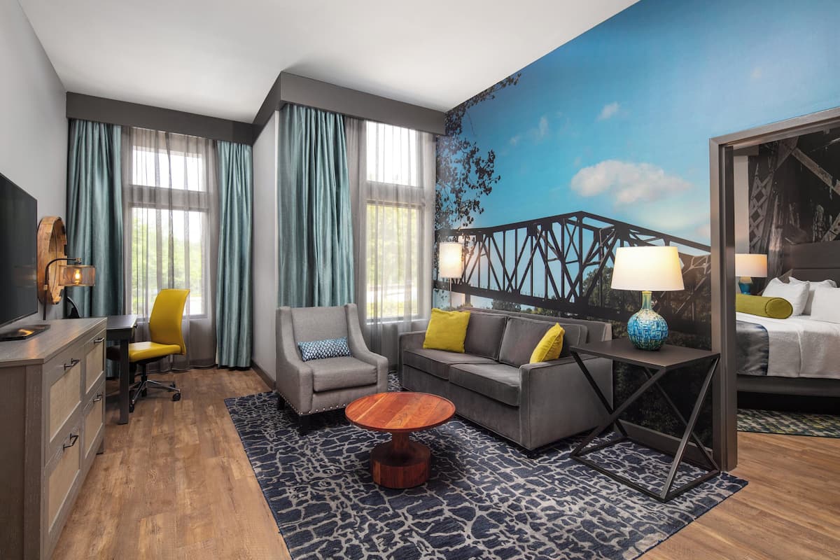 A hotel room with a modern design, featuring a sofa, armchair, and a small round table. There is a bed with a headboard, a desk with a chair, and a television. The room has large windows with curtains, a patterned rug, and a mural on the wall.