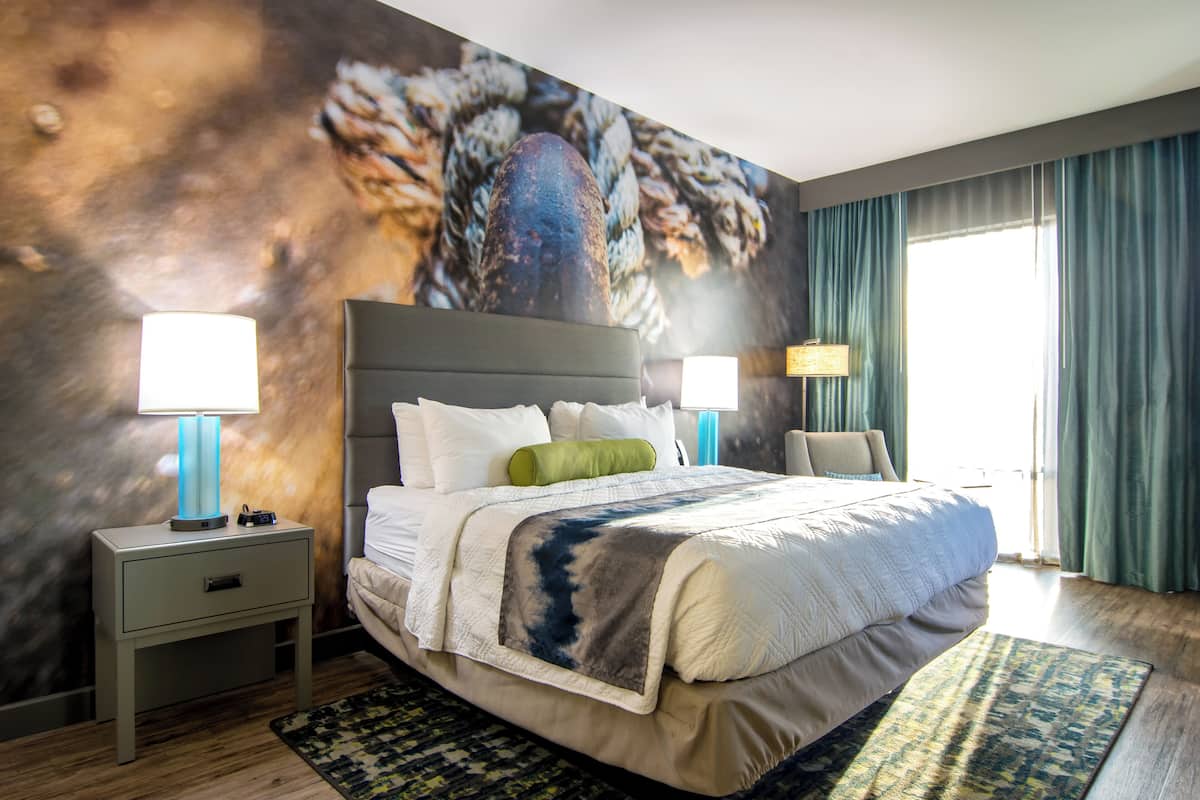 A hotel room with a large bed, two bedside tables with lamps, a green throw pillow, and a large window with curtains.