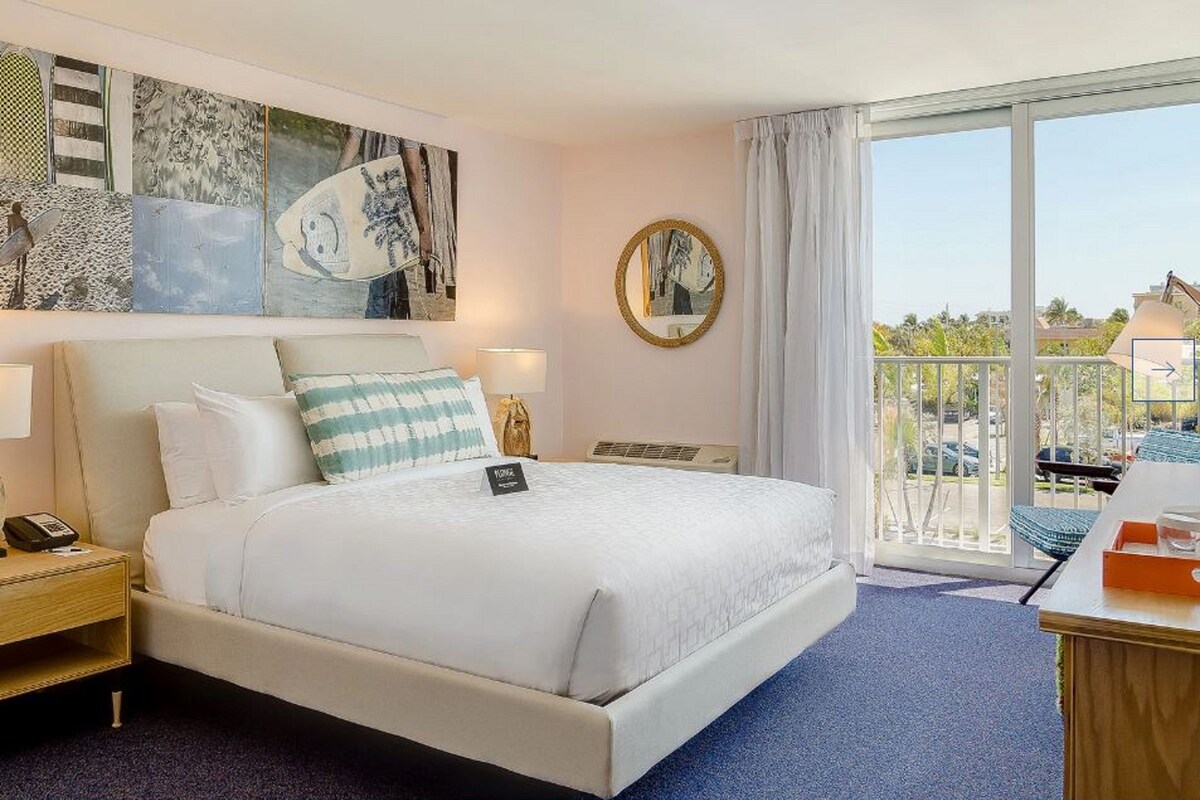 A hotel room with a bed, a nightstand, a desk, a chair, a balcony with a view, and artwork on the walls.