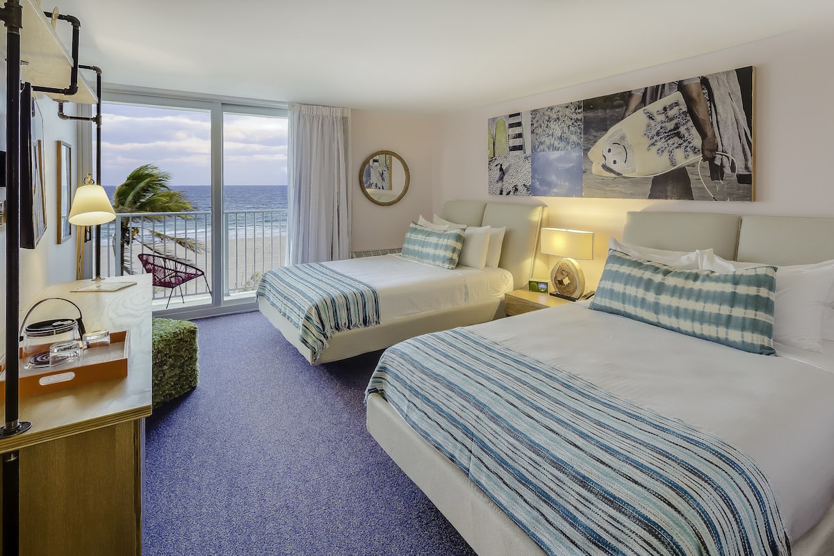 A hotel room with two beds, a desk with a lamp, a chair, and a view of the ocean.