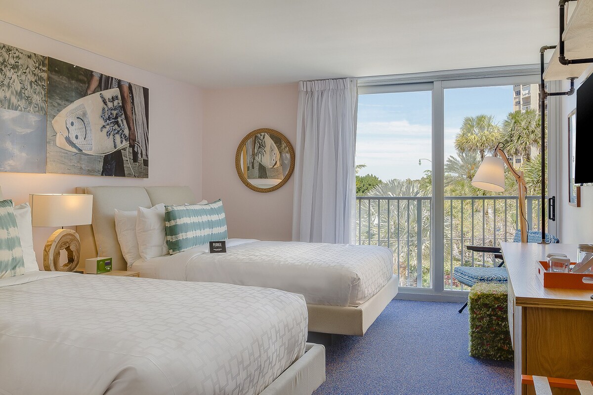 A hotel room with two beds, a balcony with a view, a desk with a lamp, and a TV.