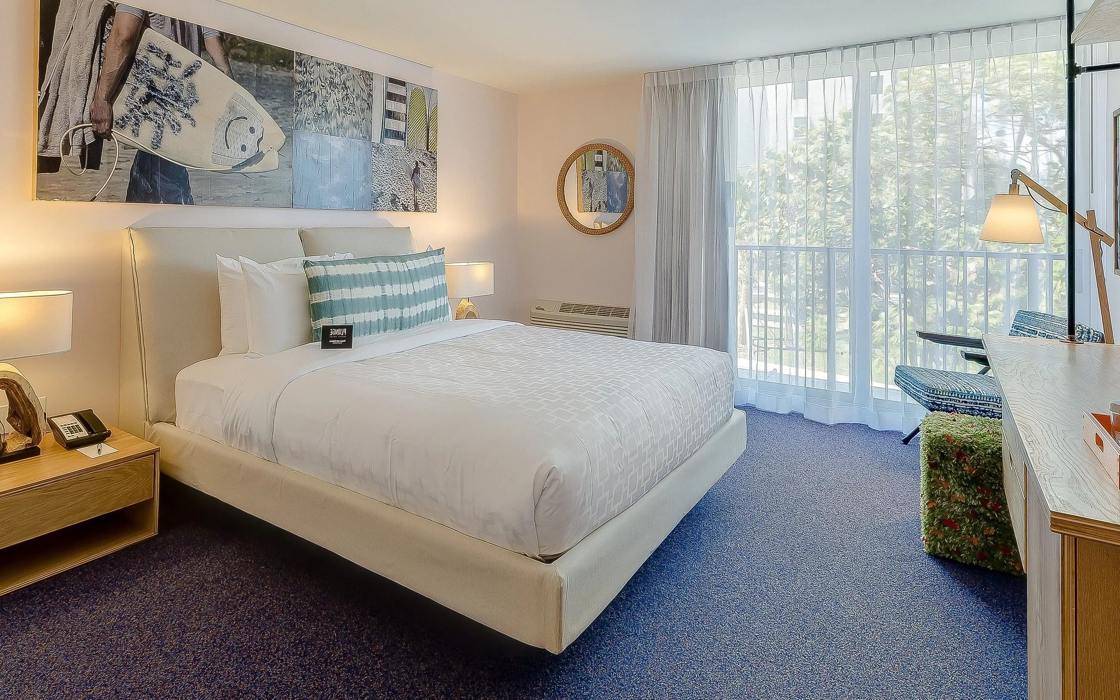 Habitación de hotel con una cama grande, una mesita de noche con lámpara, un escritorio con lámpara, un ventanal con cortinas y un cuadro en la pared.