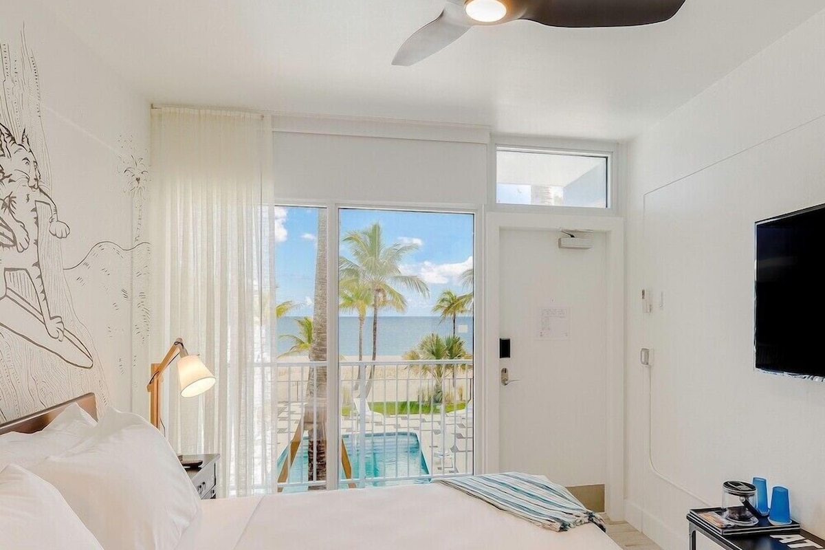 A hotel room with a bed, a ceiling fan, a TV, a balcony with a pool view, and a door.