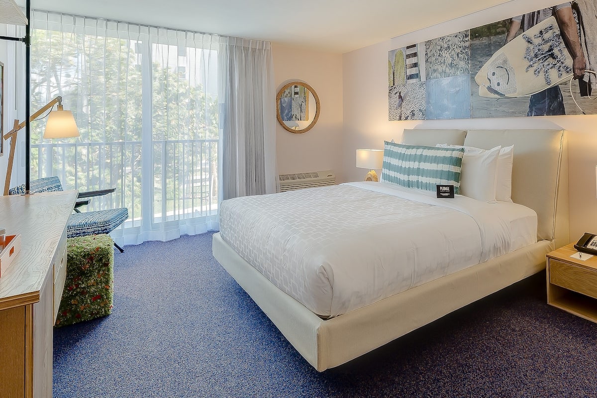 A hotel room with a large bed, a desk, a chair, a window with curtains, and a wall with artwork.