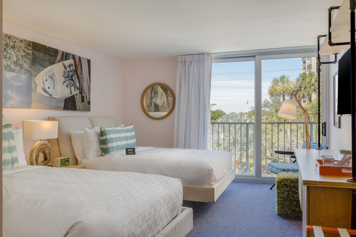 A hotel room with two beds, a balcony with a view, a desk with a lamp, and artwork on the wall.