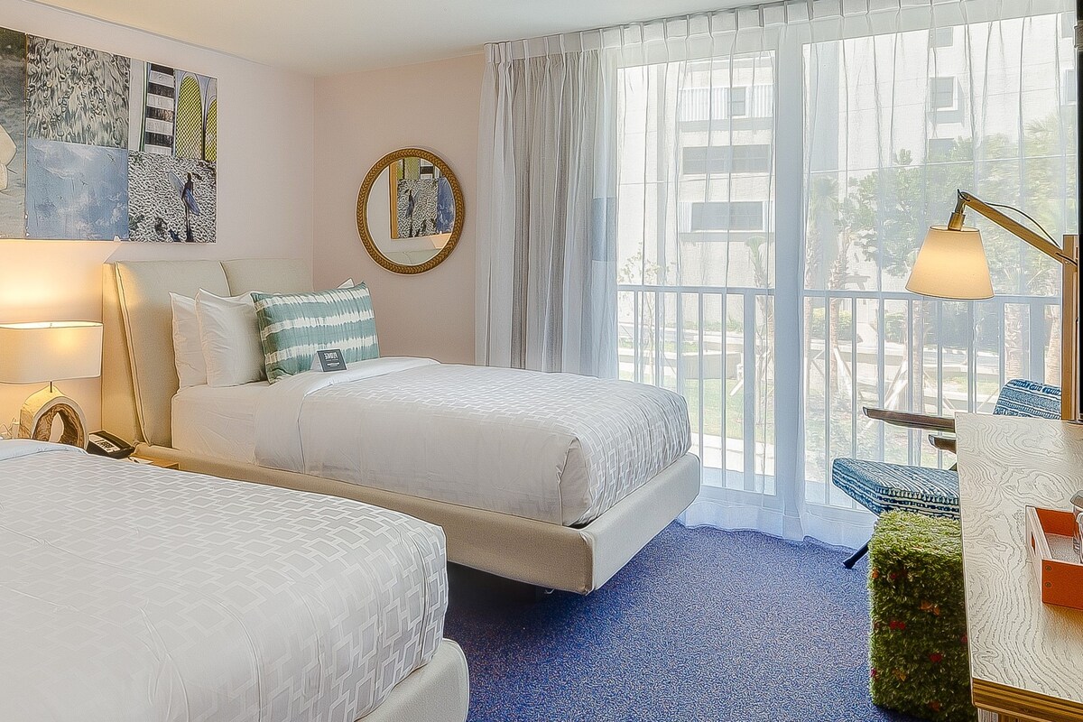 A hotel room with two beds, a large window with curtains, a round mirror, and artwork on the wall.