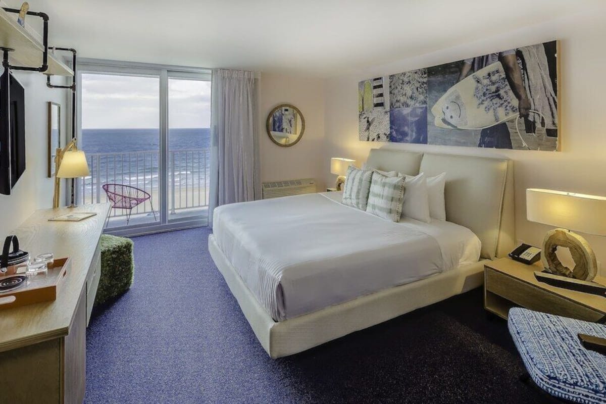 Una habitación de hotel con cama, escritorio, silla, mesita de noche, lámpara, un cuadro y vista al océano.