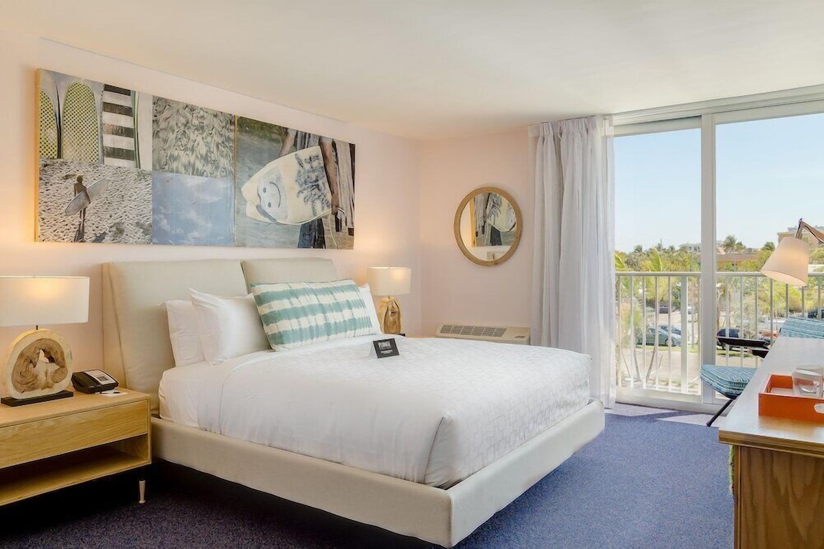 A hotel room with a bed, a nightstand, a desk, a chair, a balcony with a view, and artwork on the wall.