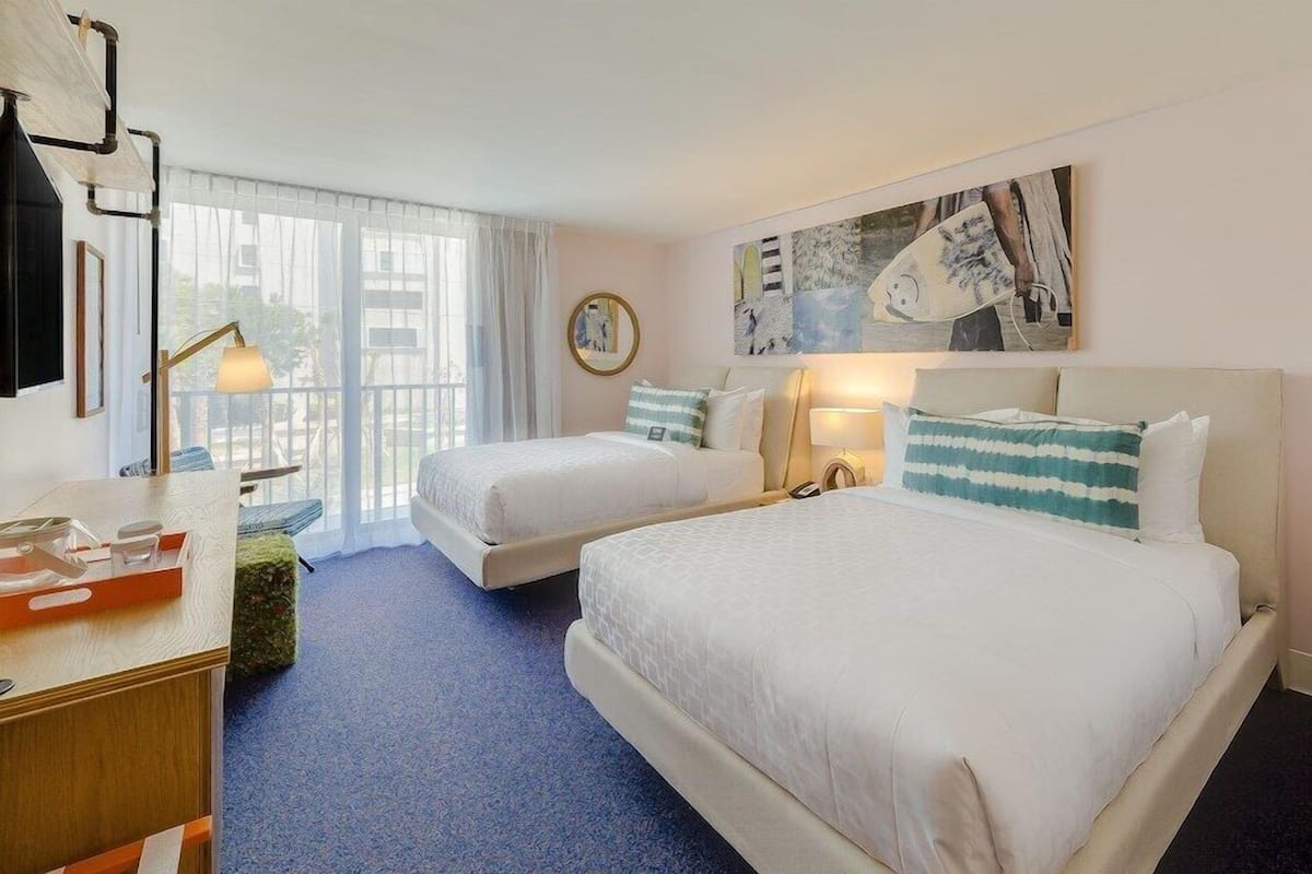 A hotel room with two beds, a desk, a chair, a window with curtains, and artwork on the wall.