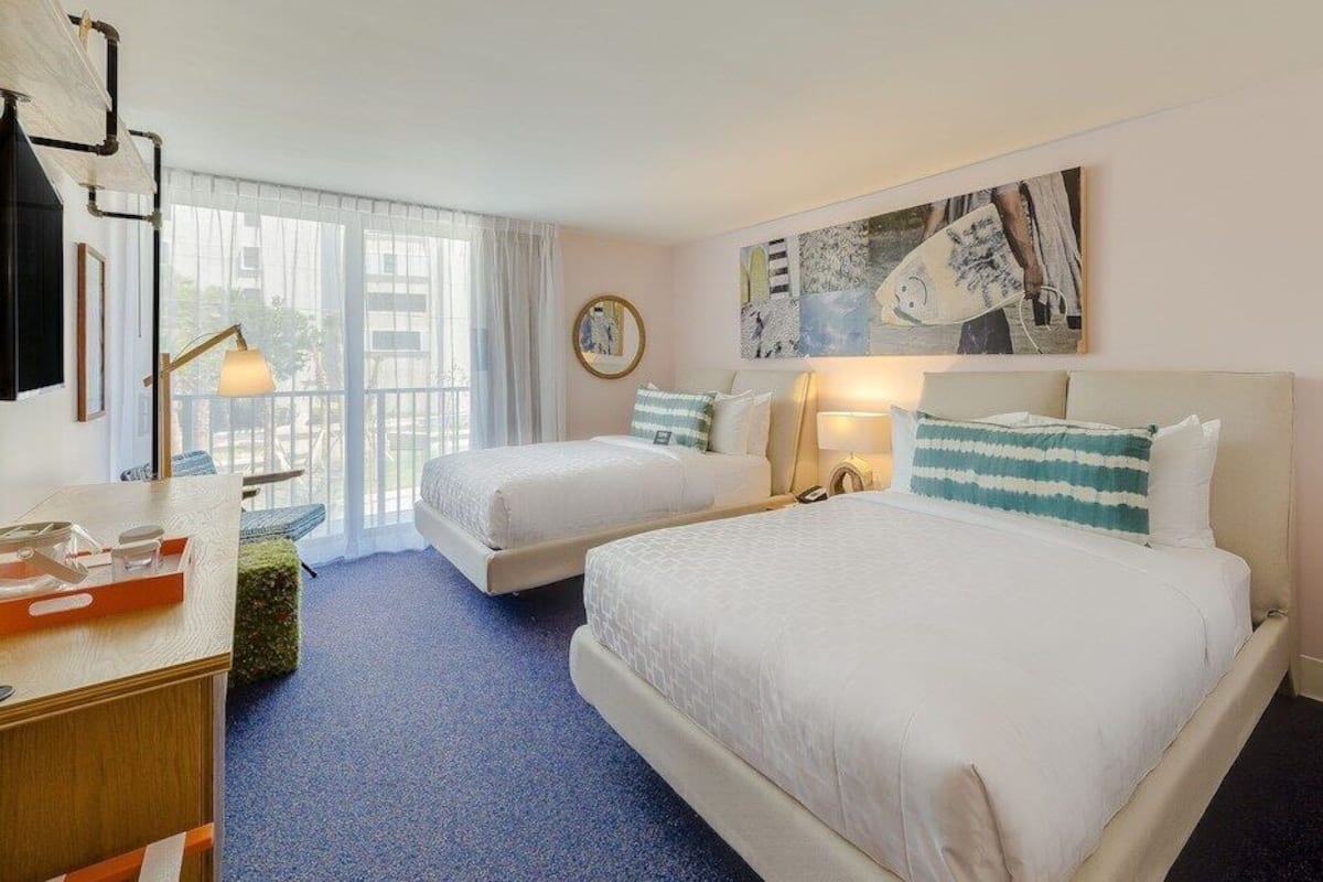 Habitación de hotel con dos camas, un escritorio, una silla, una ventana con cortinas y obras de arte en la pared.