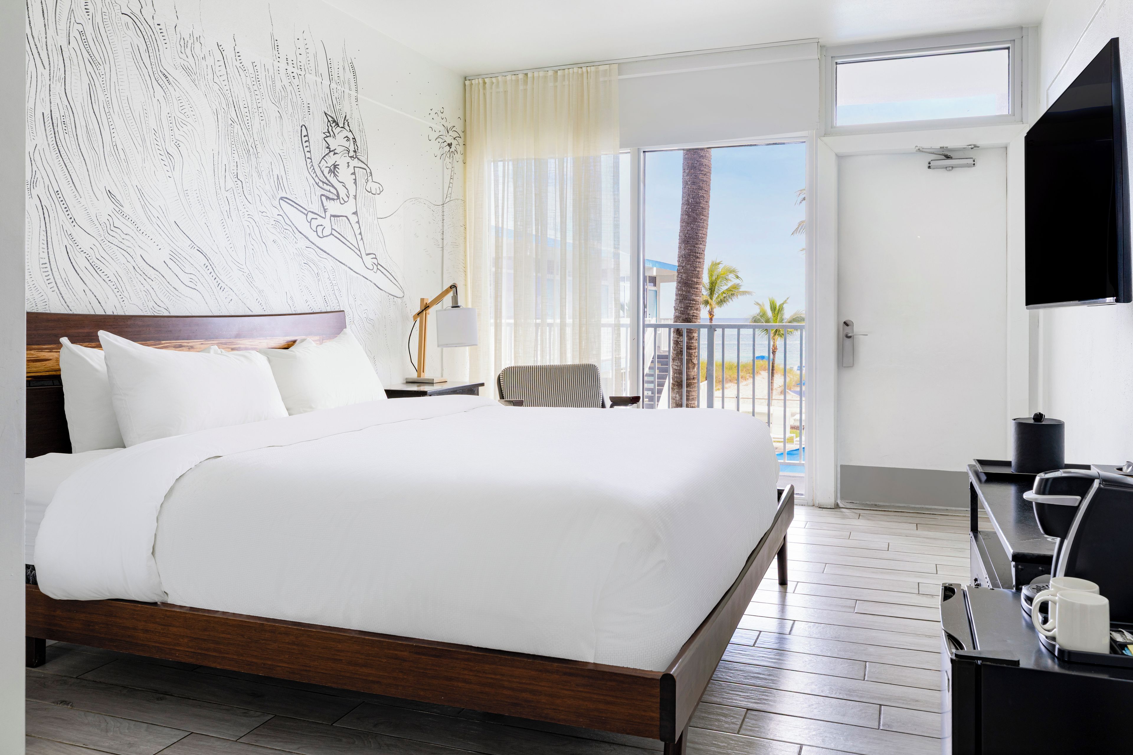 A hotel room with a large bed, a desk with a chair, a TV, and a balcony with a view of the beach.