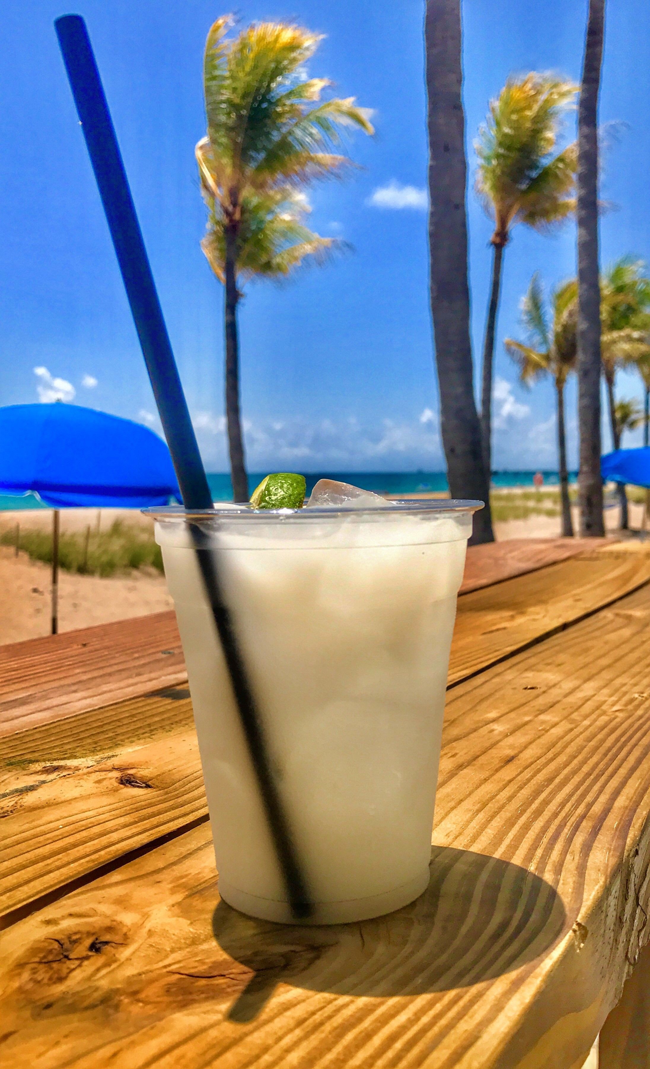 A clear glass of a beverage with a straw and lime on a wooden table at a beach with palm trees and a blue umbrella in the background.