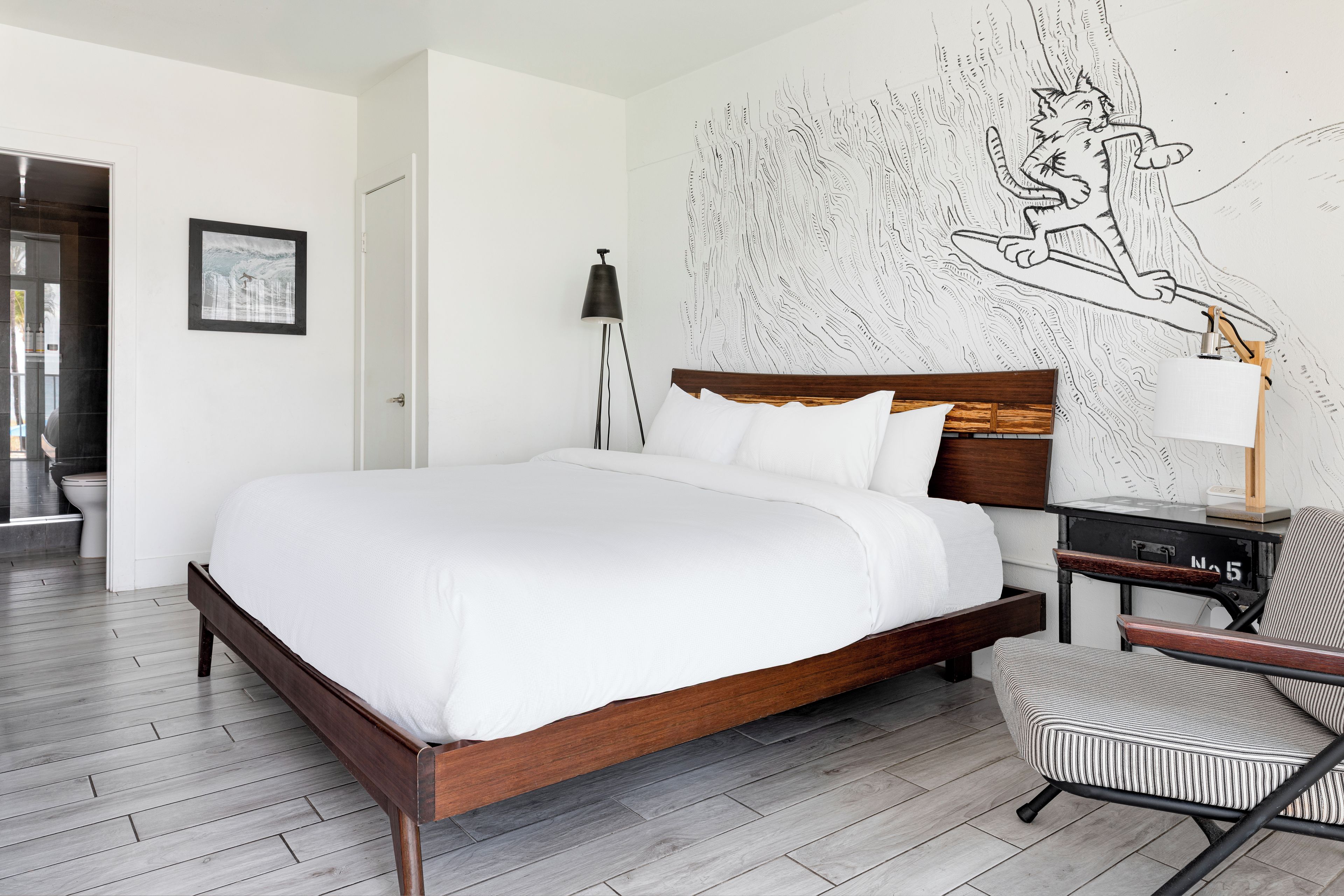 Une chambre moderne avec un lit en bois, une literie blanche, une fresque murale, un bureau avec une lampe et une chaise à rayures.