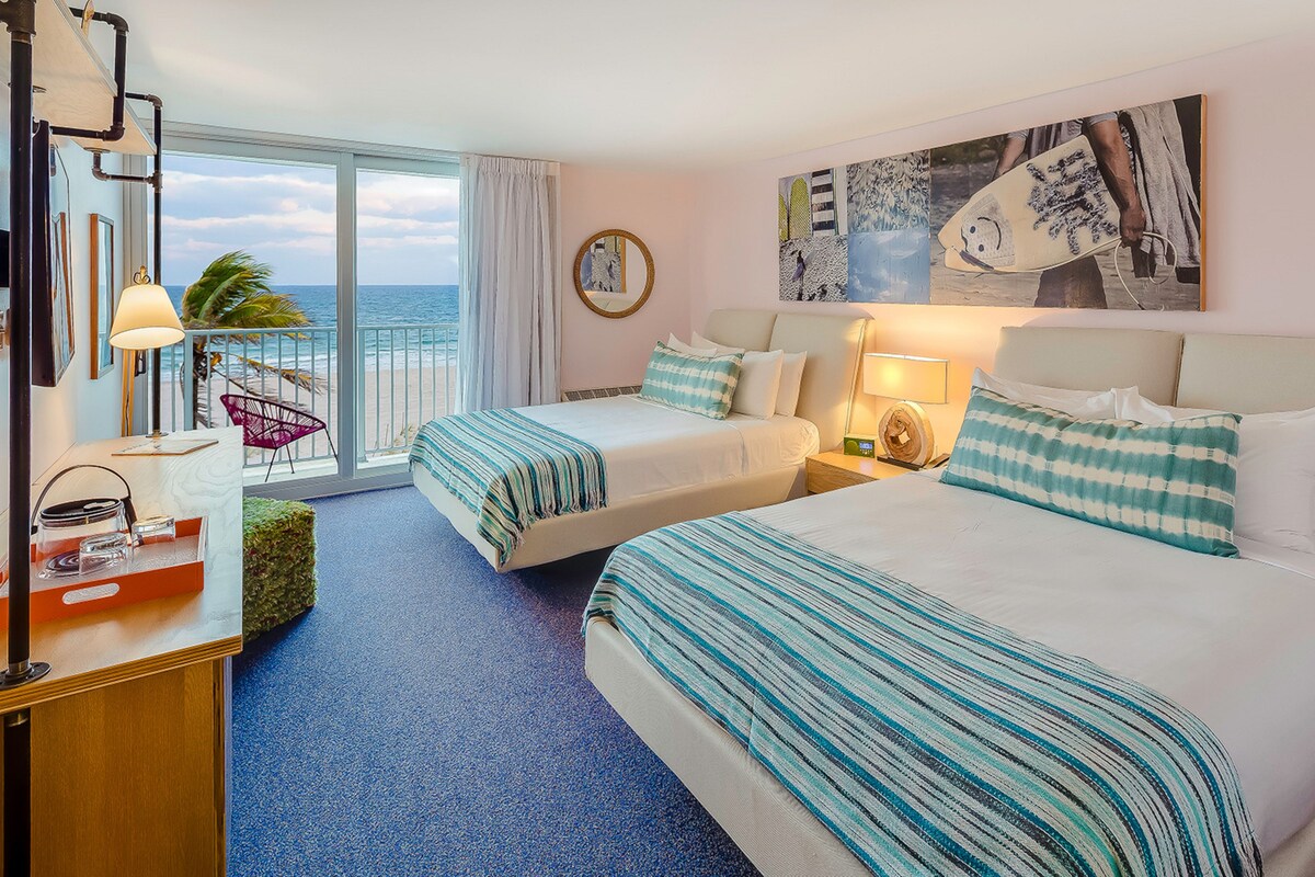 A hotel room with two beds, a desk, a chair, a view of the ocean, and artwork on the wall.