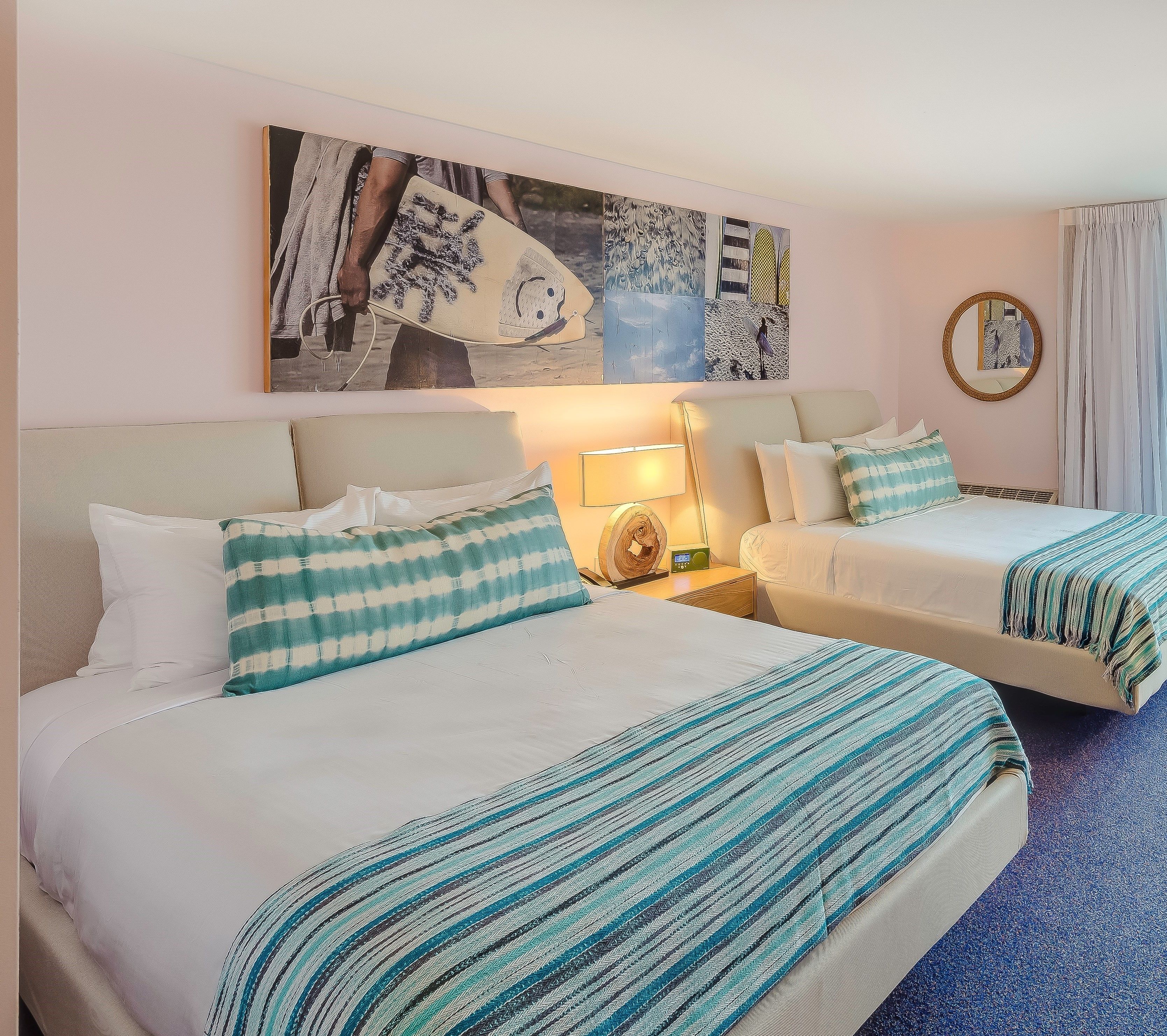 Une chambre d’hôtel avec deux lits, chacun recouvert d’une courtepointe à carreaux blancs et turquoise, assortie aux coussins. On y trouve une table de chevet avec une lampe et une petite table avec une horloge. Une grande peinture abstraite est accrochée au-dessus des lits.