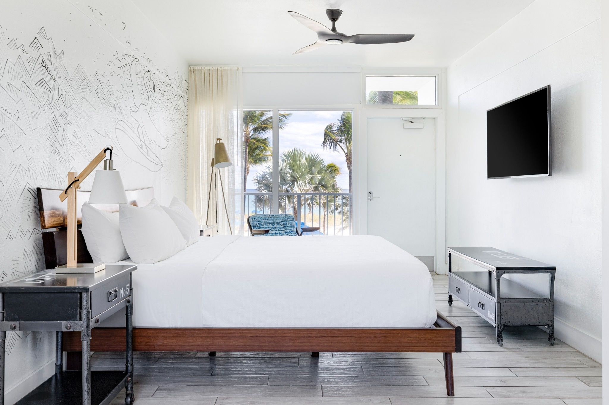 A bedroom with a bed, a desk, a chair, a TV, and a balcony with a view of palm trees.