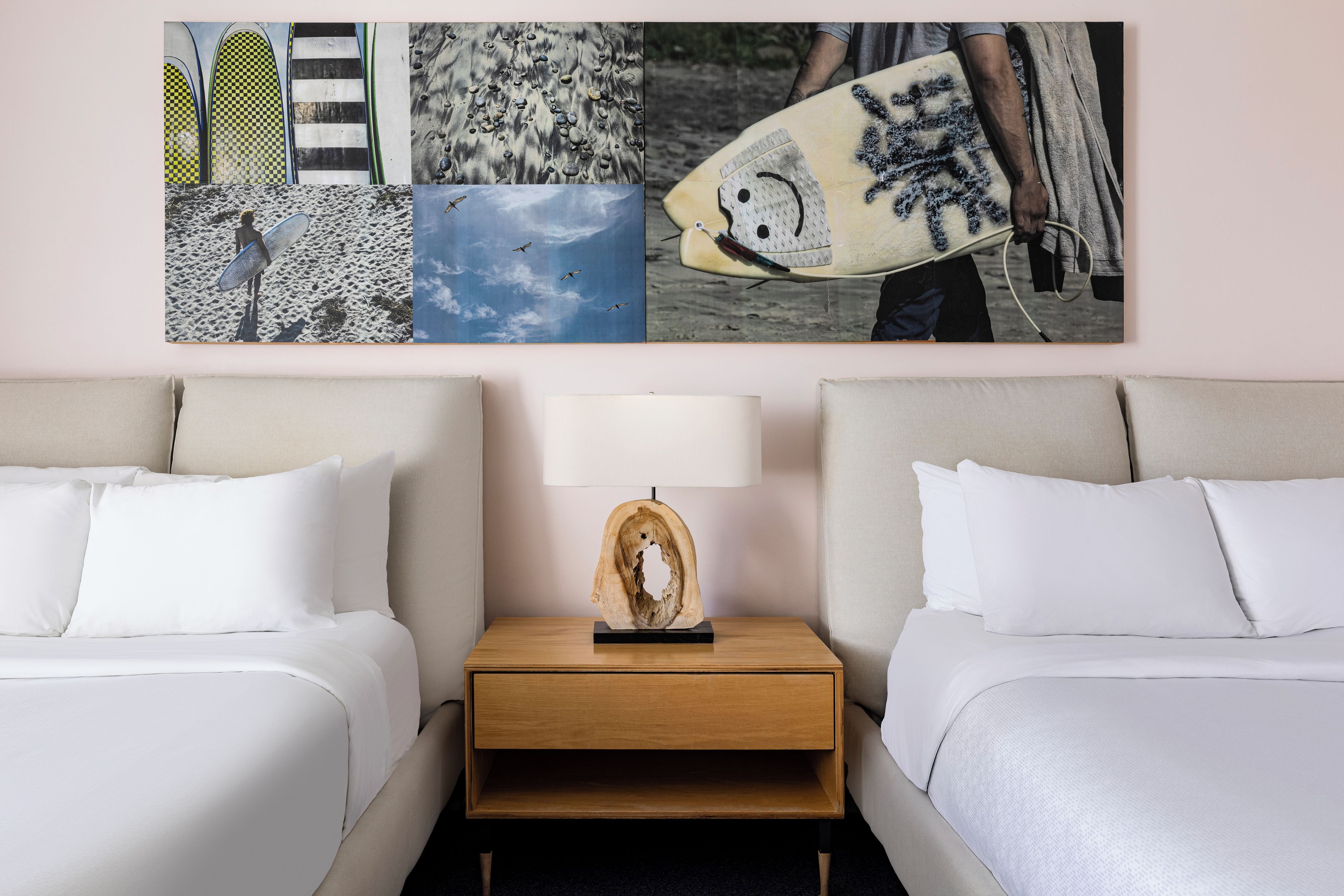 A hotel room with two beds, a nightstand with a lamp, and a wall with a collage of surfboards and beach scenes.