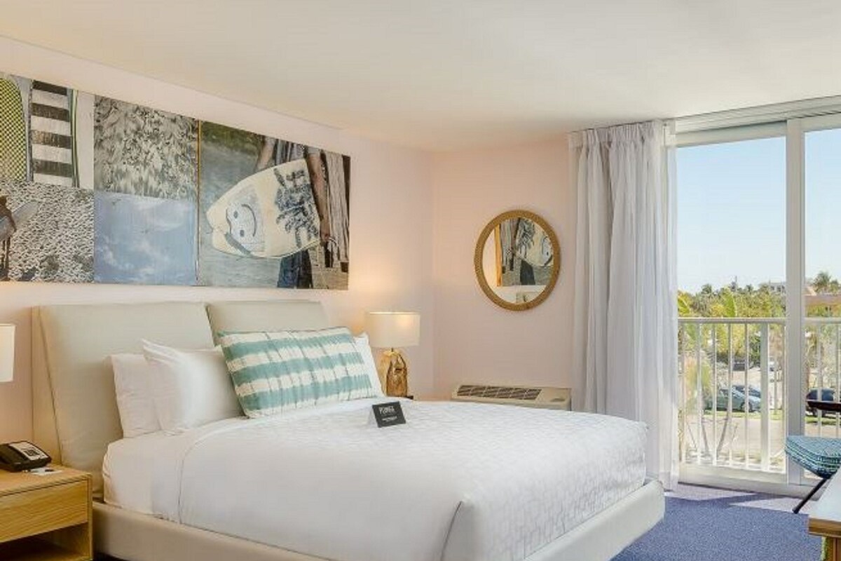 A hotel room with a bed, a nightstand, a lamp, a round mirror, and artwork on the wall.