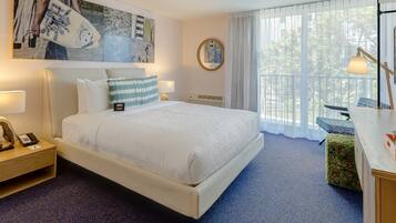 A hotel room with a large bed, a nightstand with a lamp, a desk with a lamp, a large window with a view of greenery, and a wall with a framed artwork.