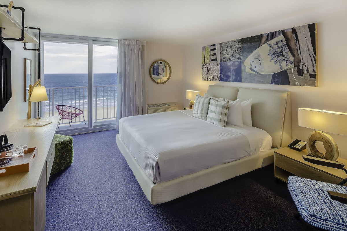 A hotel room with a bed, a desk, a chair, a lamp, a phone, and a painting.