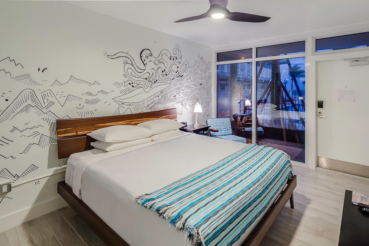 A hotel room with a bed, a desk with a lamp, a chair, and a view of the outside.