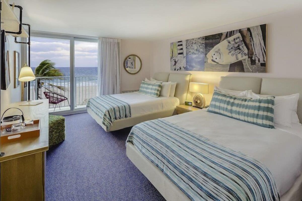 A hotel room with two beds, a desk, a chair, and a view of the ocean.