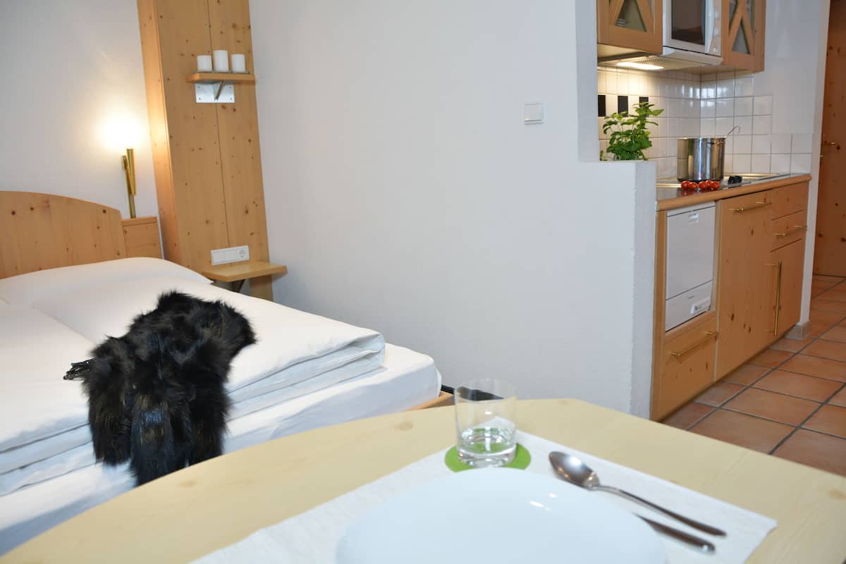 Habitación de hotel con cama, mesita de noche con un vaso de agua, plato con cuchara y área de kitchenette con armarios y microondas.