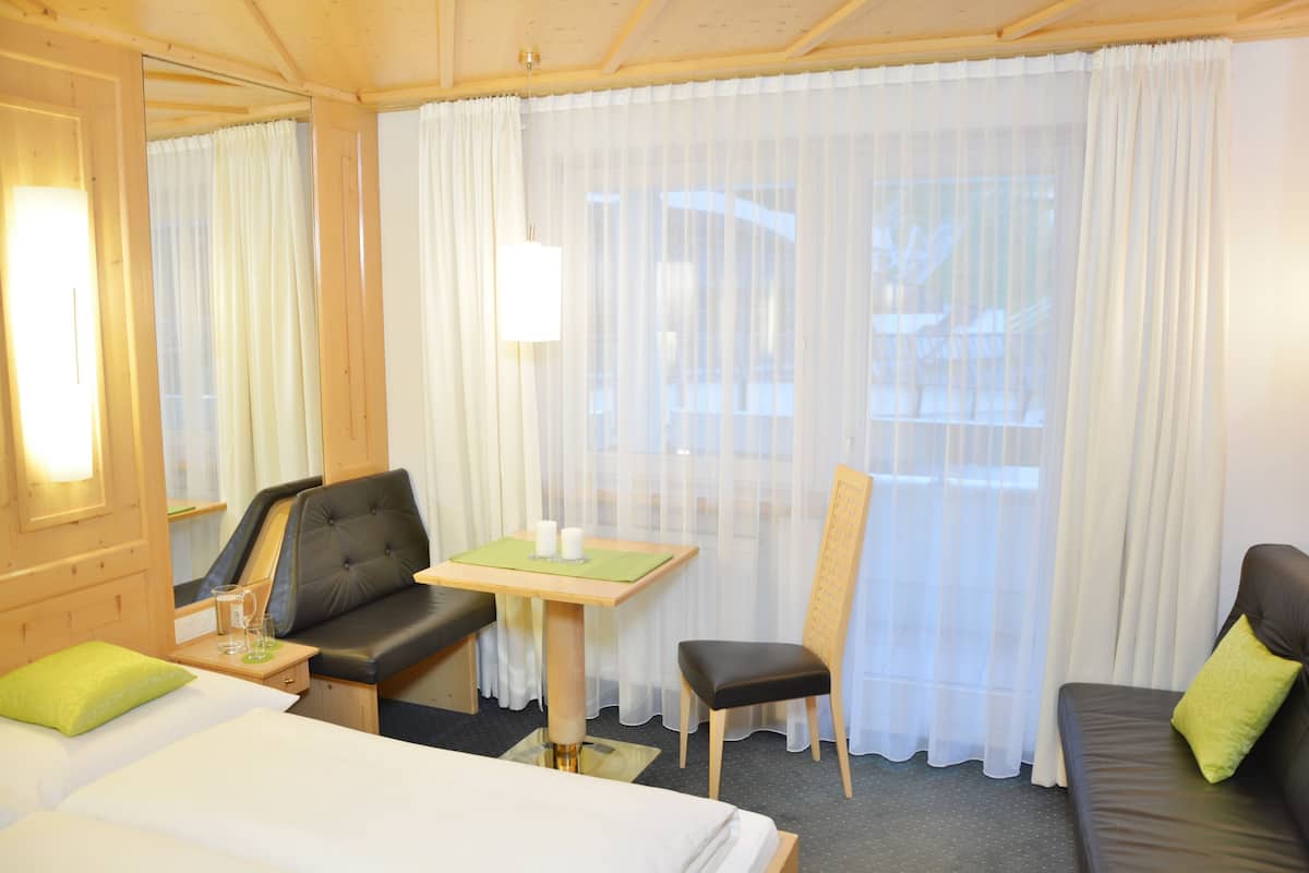 Una habitación de hotel con una cama, un escritorio con una silla, un sofá y un ventanal con cortinas traslúcidas.