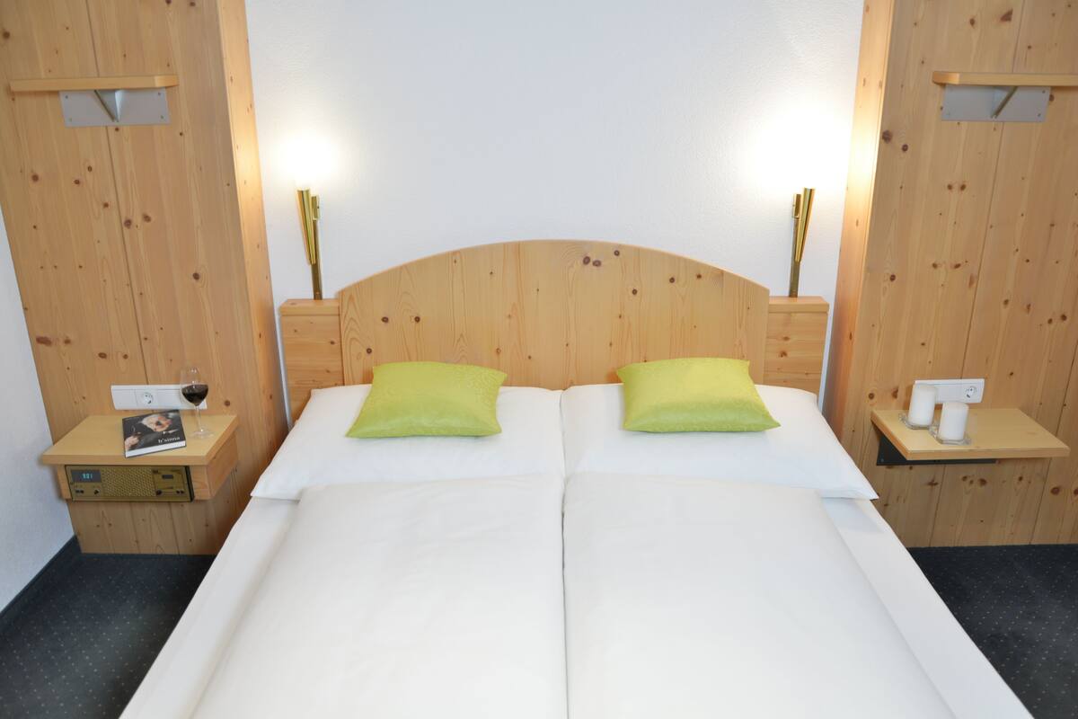 Cama doble con dos almohadas, cabecera de madera, mesitas de noche con lámparas, y una mesita auxiliar con un libro y una copa de vino.