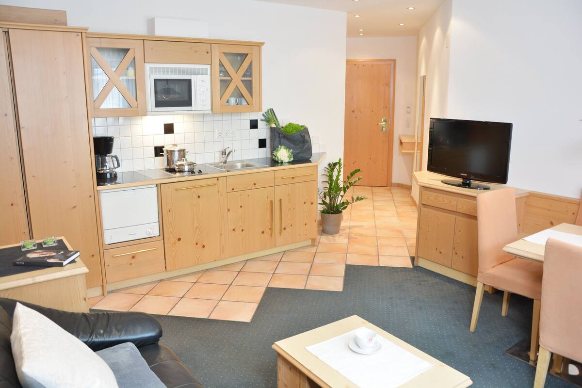 Una cocina moderna con armarios de madera, microondas y cafetera. Hay una zona de comedor con mesa y sillas, y un televisor sobre un soporte.