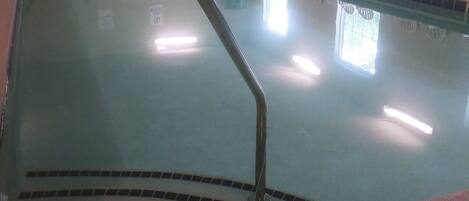 An indoor swimming pool with a tiled edge, a metal handrail, and white chairs arranged around the pool.