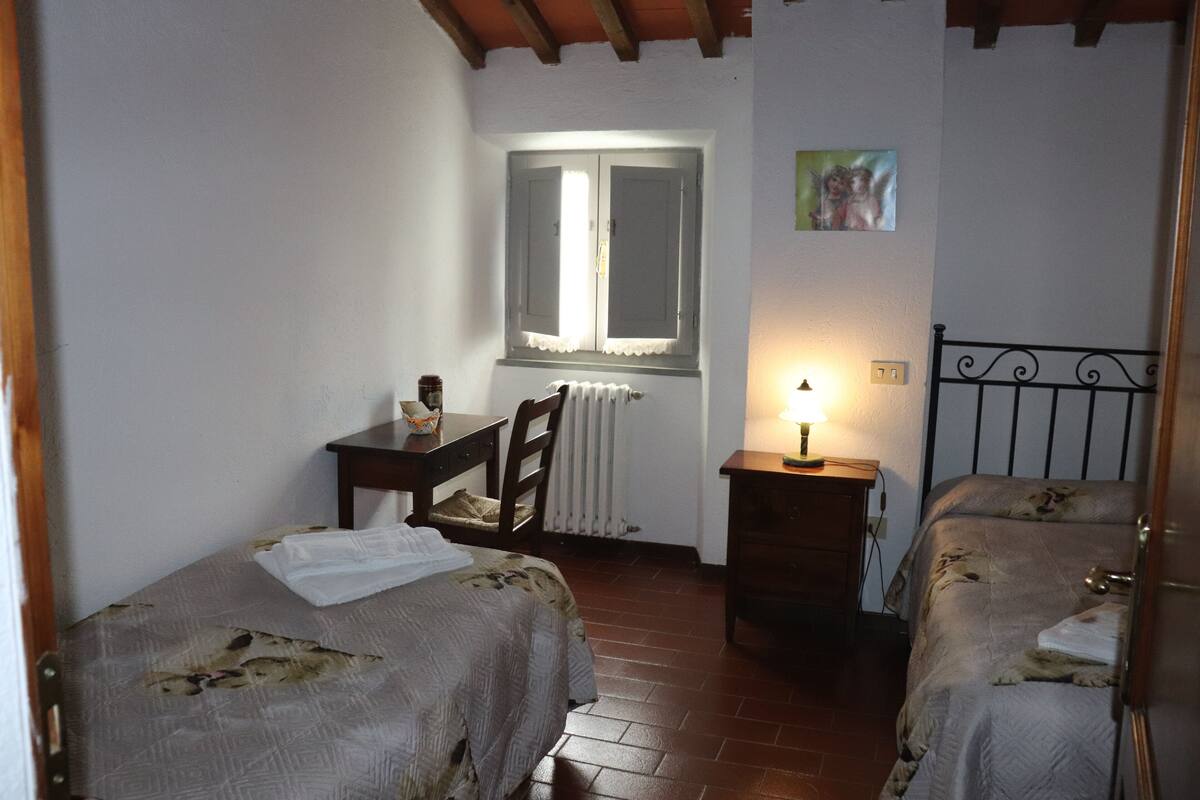 A bedroom with two beds, a desk, a chair, a radiator, and a window with shutters.