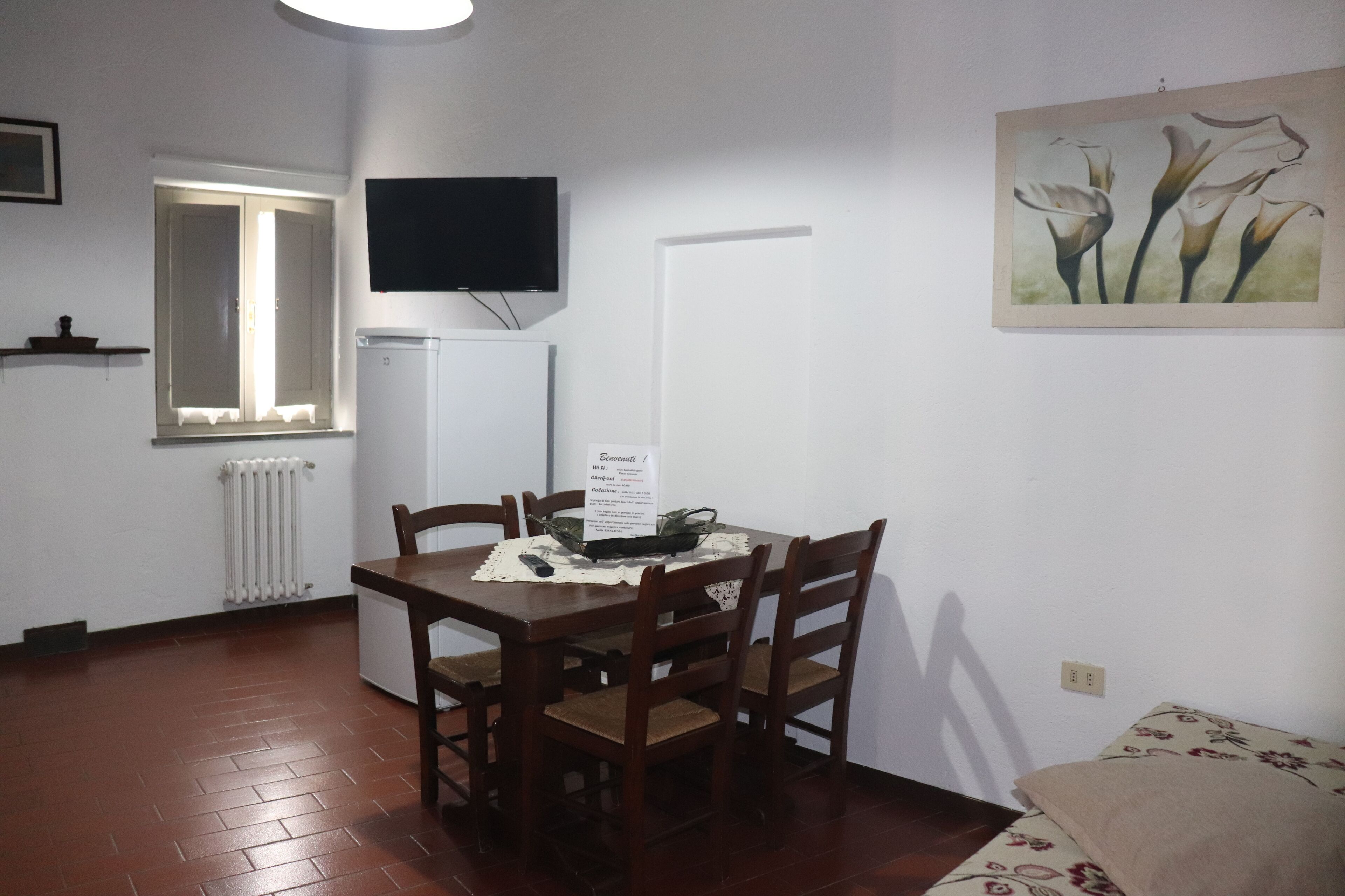Zona pranzo con tavolo, sedie, televisione a parete, termosifone e finestra con tende.