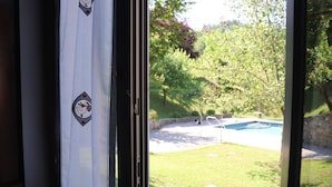 A view through a window with a curtain featuring a pattern of birds. Outside, there is a swimming pool surrounded by a grassy area and trees.