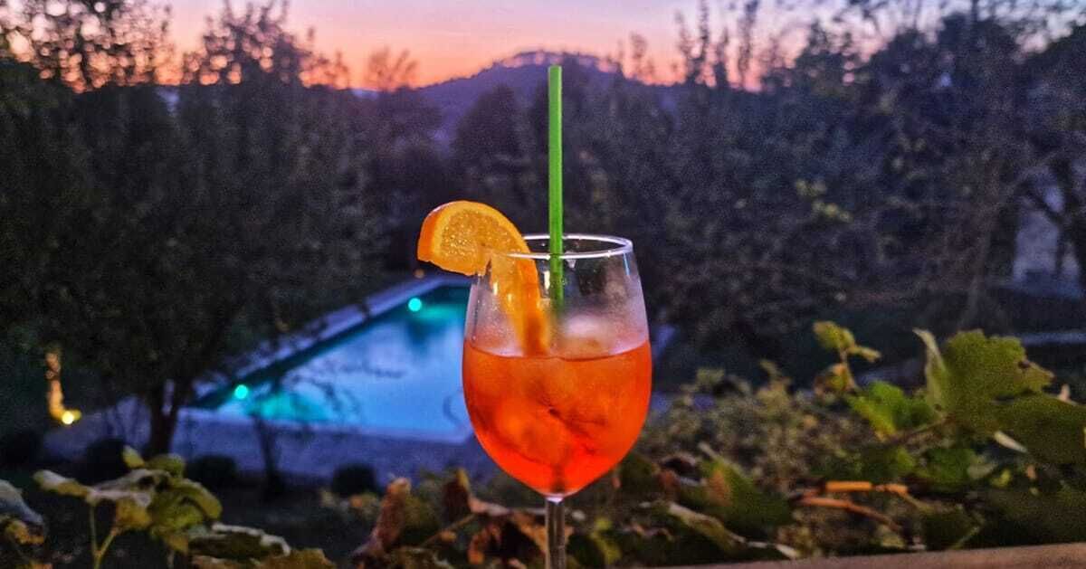 A glass of red beverage with an orange slice garnish, placed outdoors with a view of a swimming pool and trees in the background.